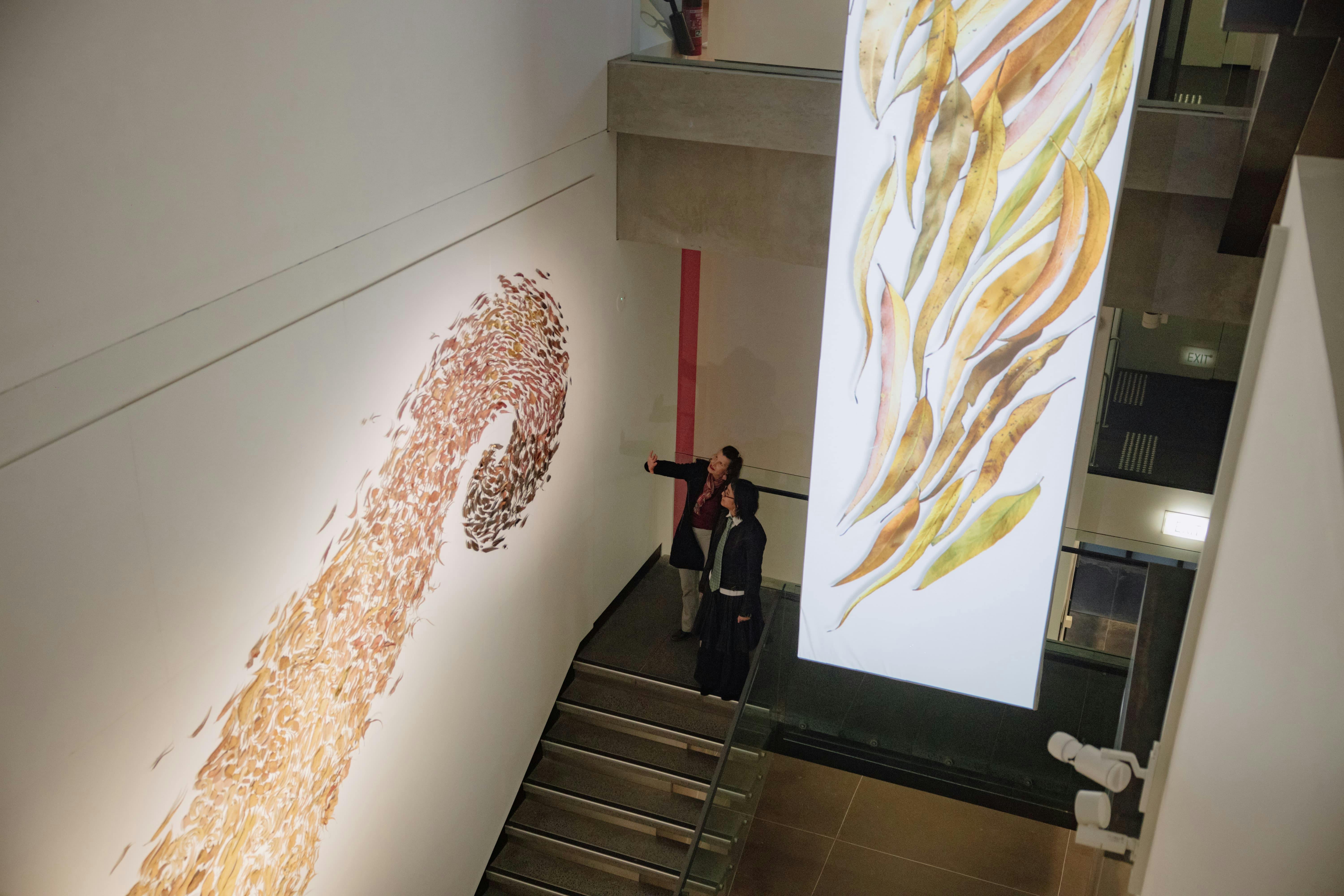 Two people on a staircase, looking up at an artwork of leaves on a wall and a leaves projection.