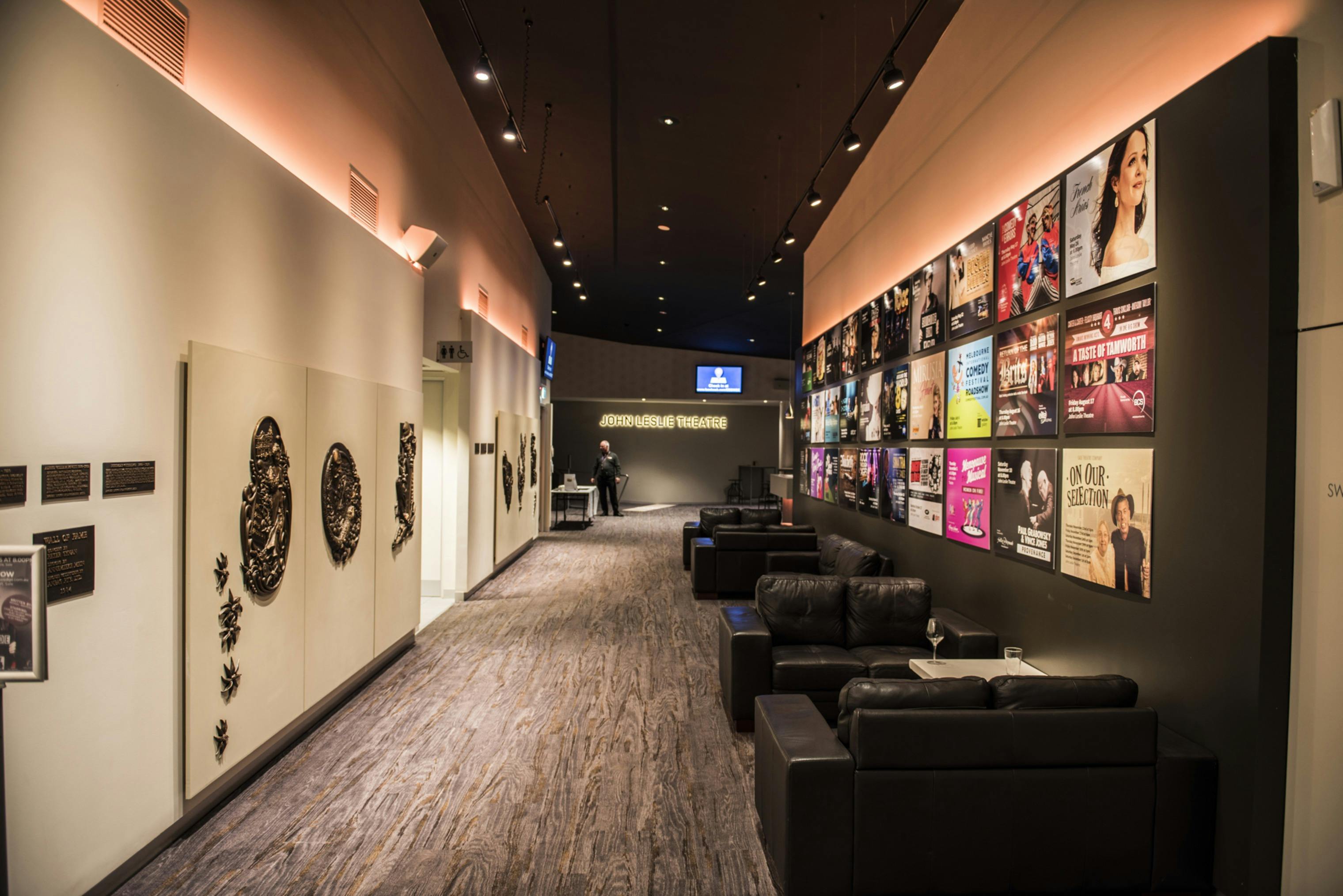 a corridor with black couches and the text John Leslie Theatre on a wall, with posters on walls