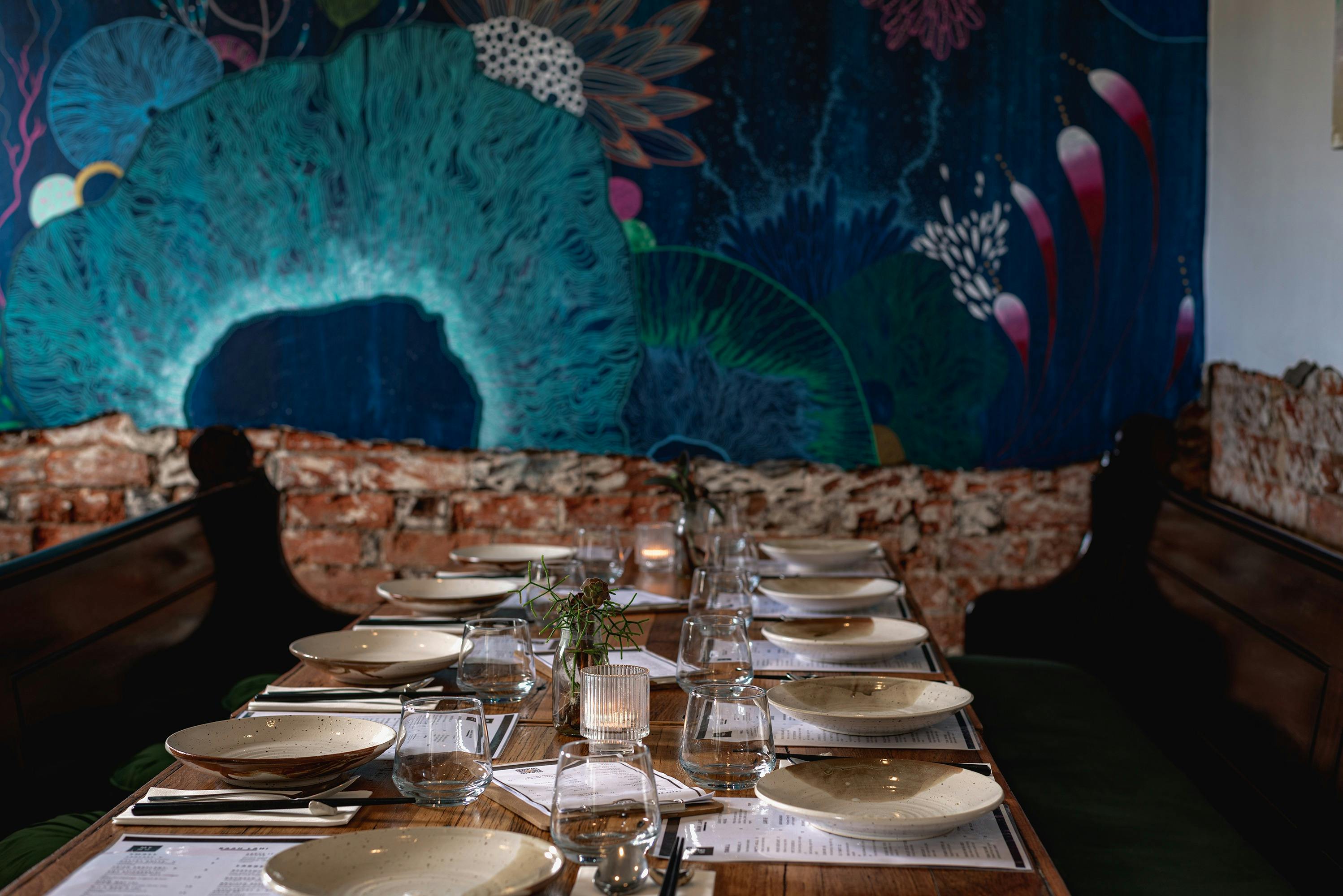 A set table with church pews on either side in front of brightly multi-coloured mural on wall