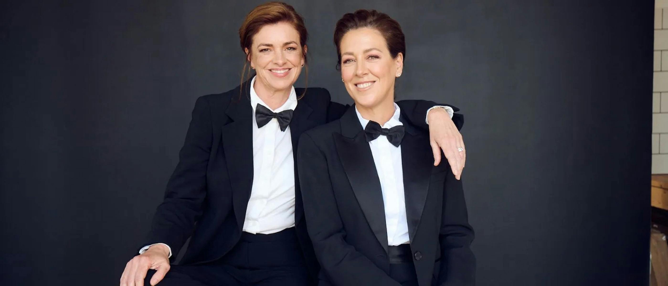 Two women wearing suits and bowties sitting together, smiling.