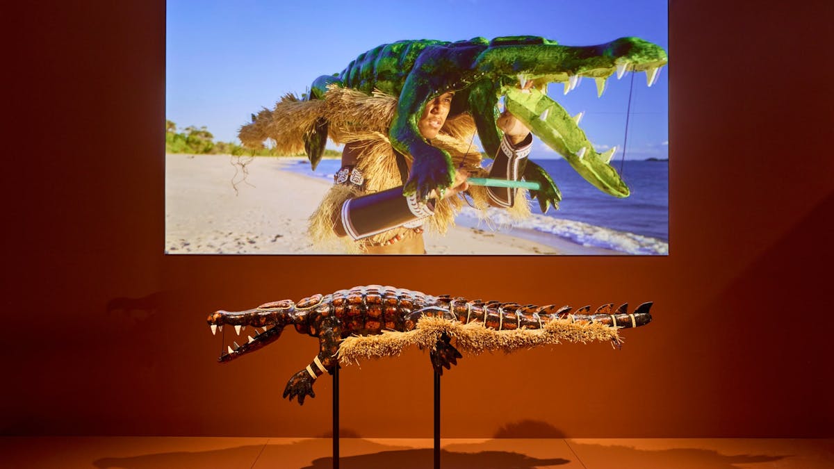 A crocodile headdress is mounted in a gallery in front of a screen displaying a video of a dancer