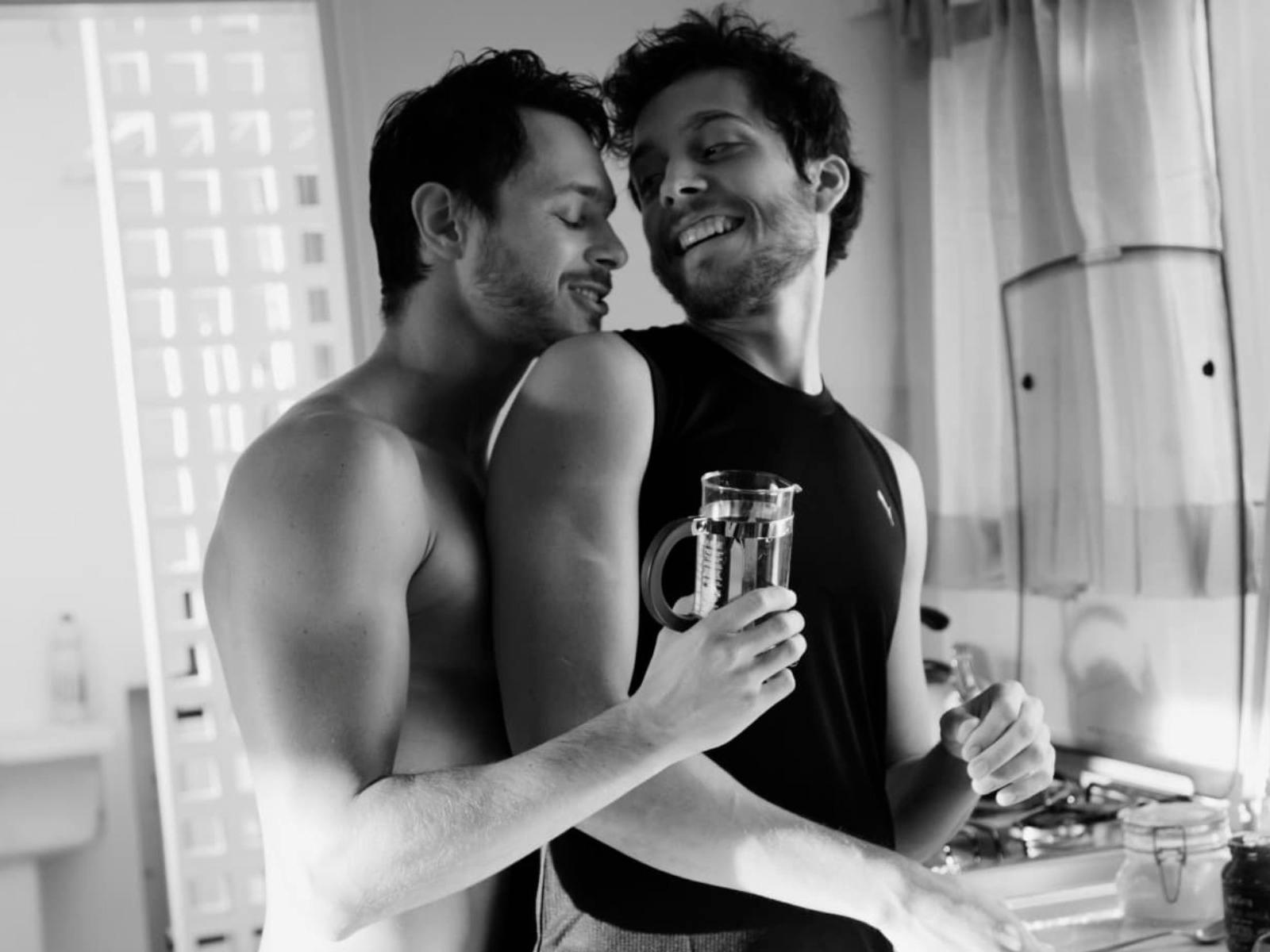 A man embracing another from the back, both smiling at each other.