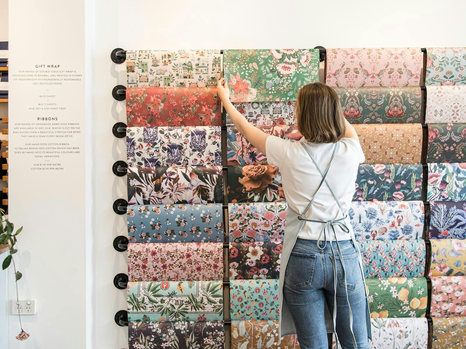Woman choosing floral gift wrap from a colourful wall display in a store.