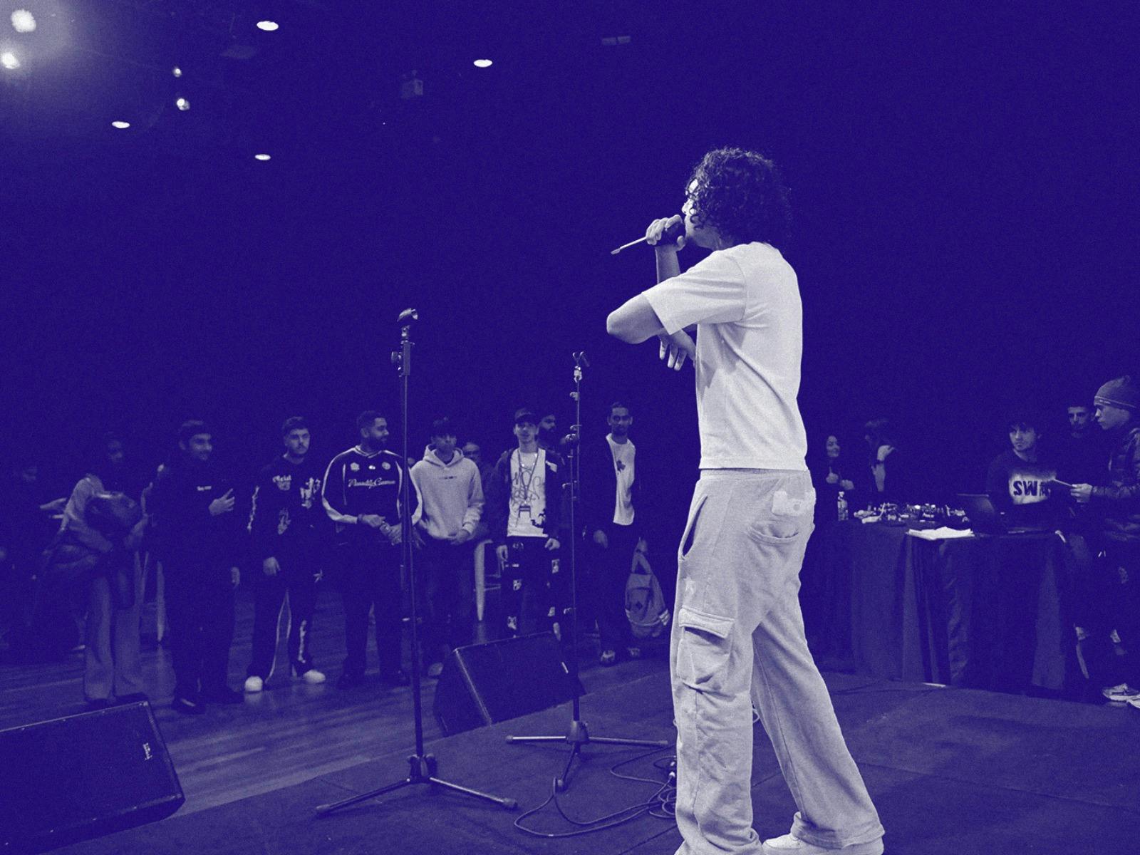 Musician singing in front of audience. Image has a purple wash over it and a grain effect