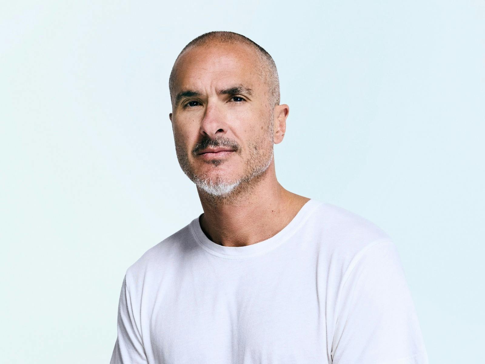 Man with shaved head and short beard wearing a white shirt looks calmly at camera.