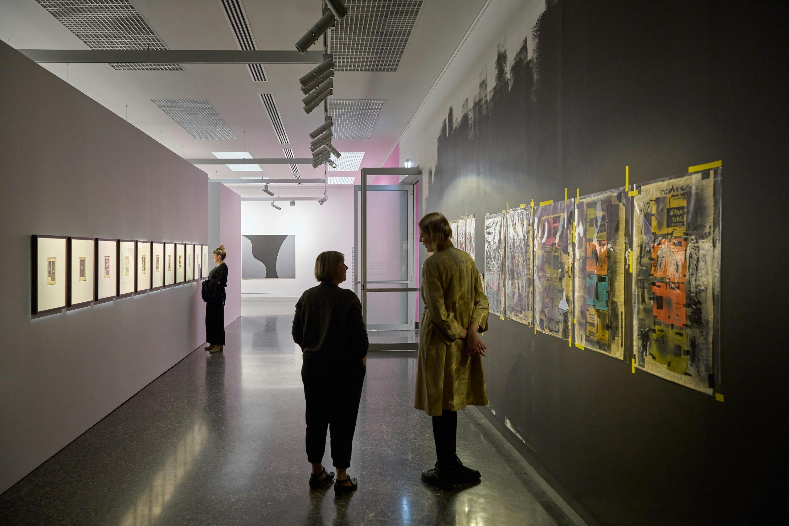 Two people stand in front of many newspaper cutouts are stuck across darkly painted wall