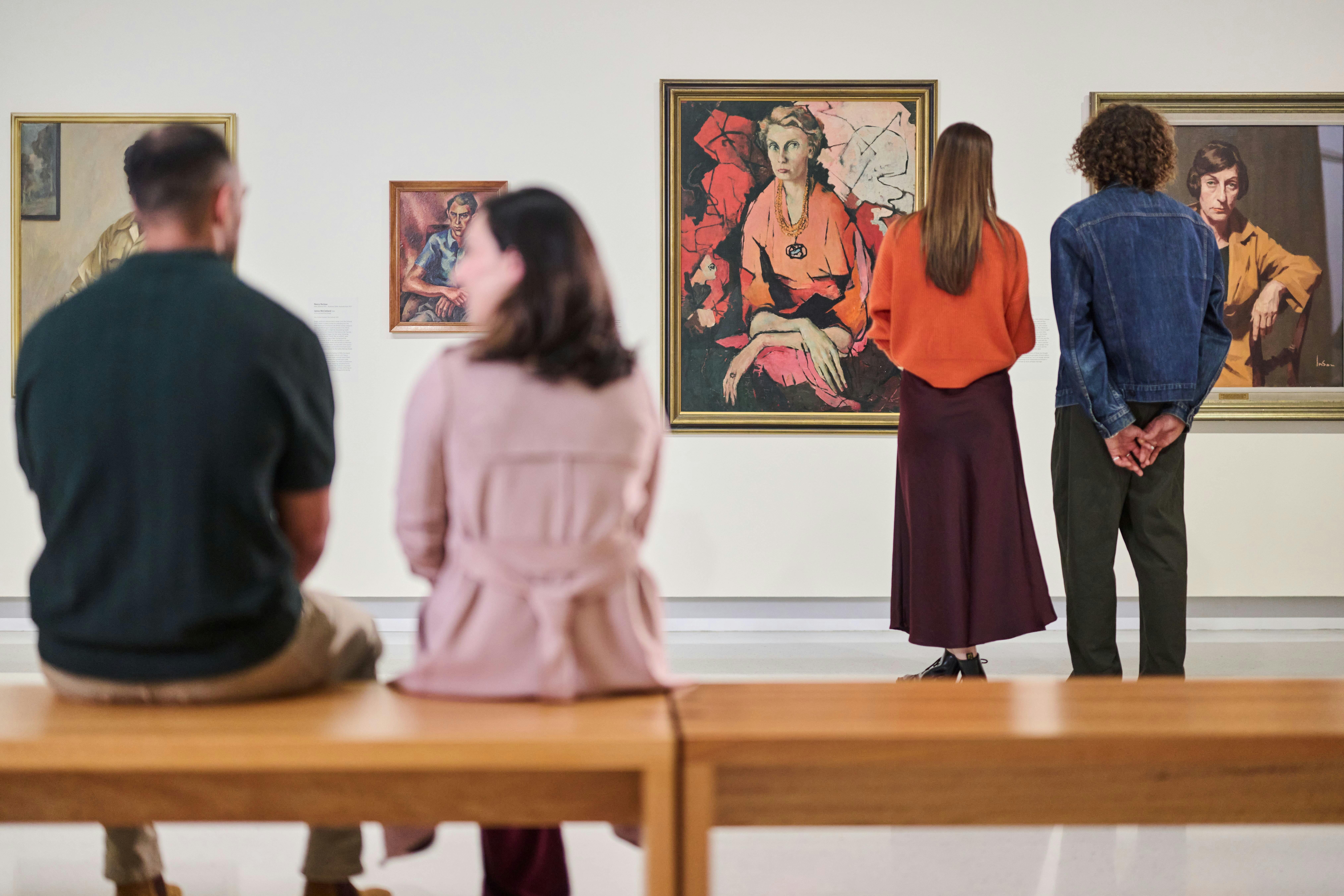 Visitors look at painted portraits in a Gallery; in the foreground, a couple sits together.