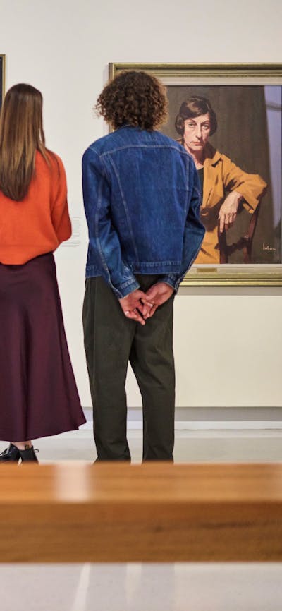 Visitors look at painted portraits in a Gallery; in the foreground, a couple sits together.