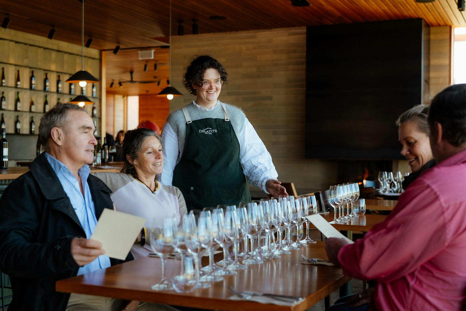 Four people sitting down with wine glasses in front of them being served by someone standing up