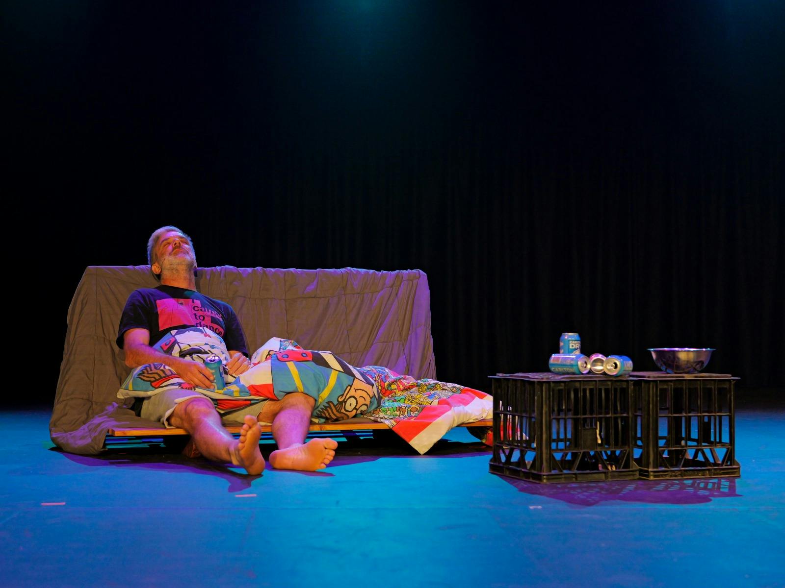 一名男子斜倚在地上，身上盖着毯子，牛奶箱上放着空饮料罐。