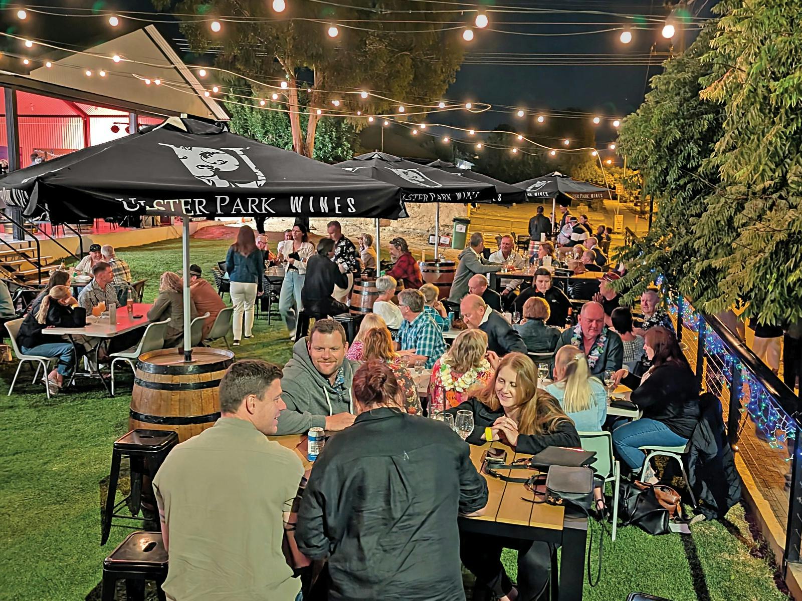 A group of people sit on the frpnt lawn of Ulster Park Wines, enjoying wine, beer, and pizza.