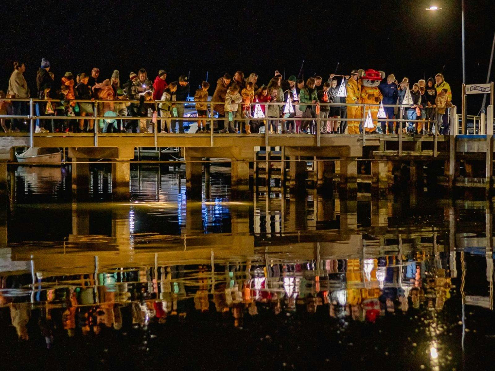 Crowd of people standing on wharf at night with glowing lanterns reflecting in water below