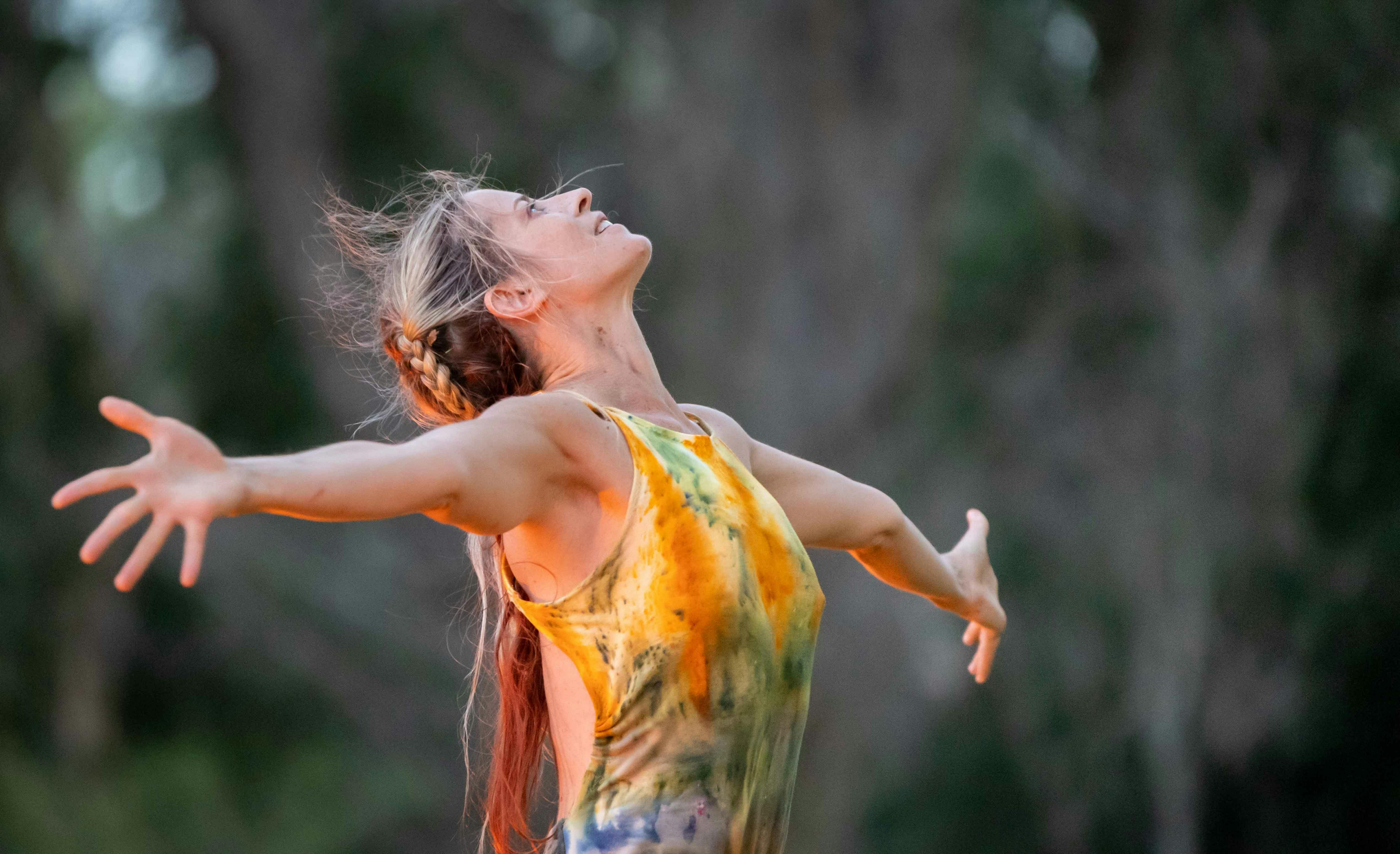 A dancer wearing a yellow dress with arms outstretched and head back in nature