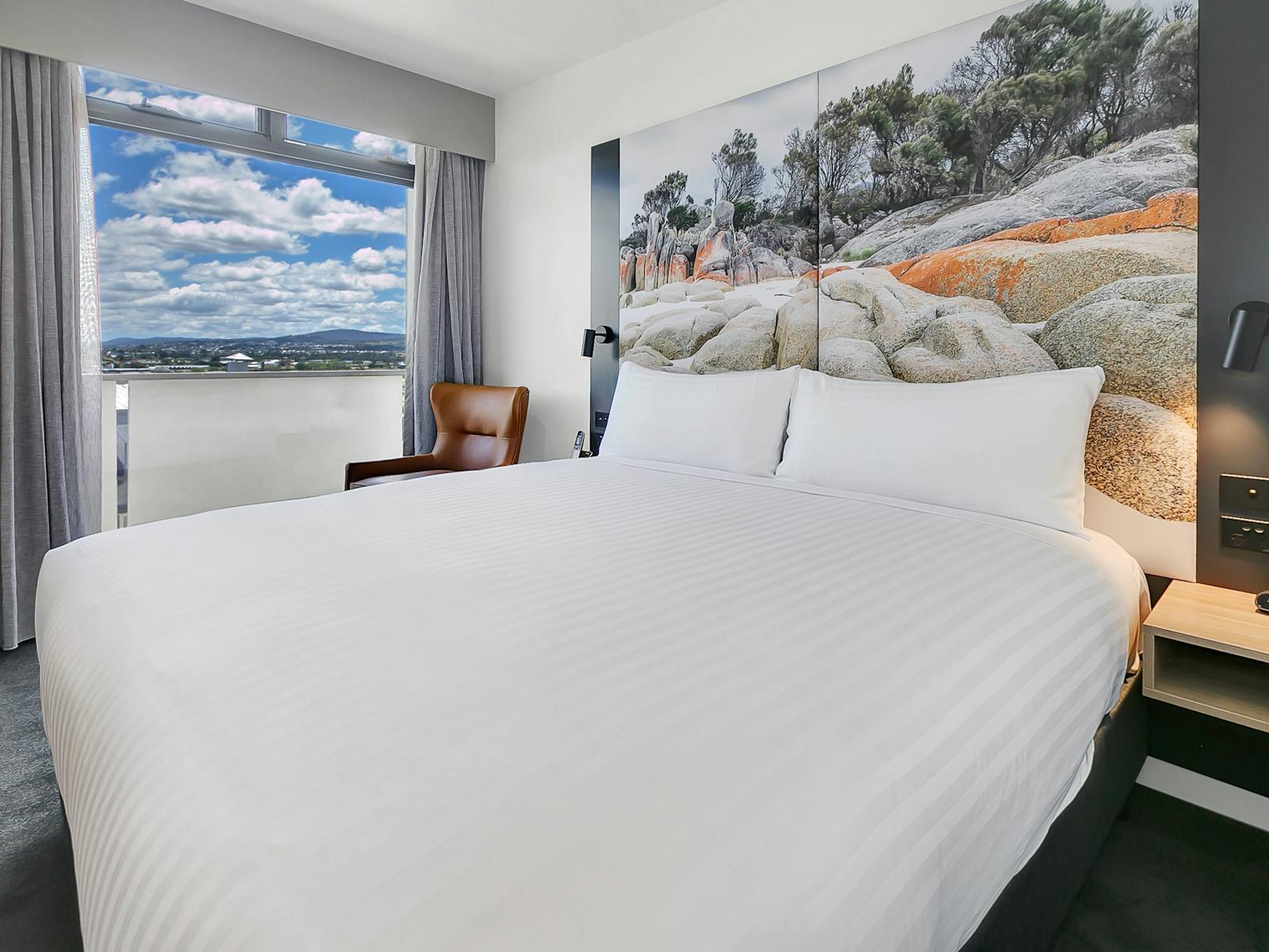 Hotel room, king bed with white linen, large picture headboard, and window with sky view.