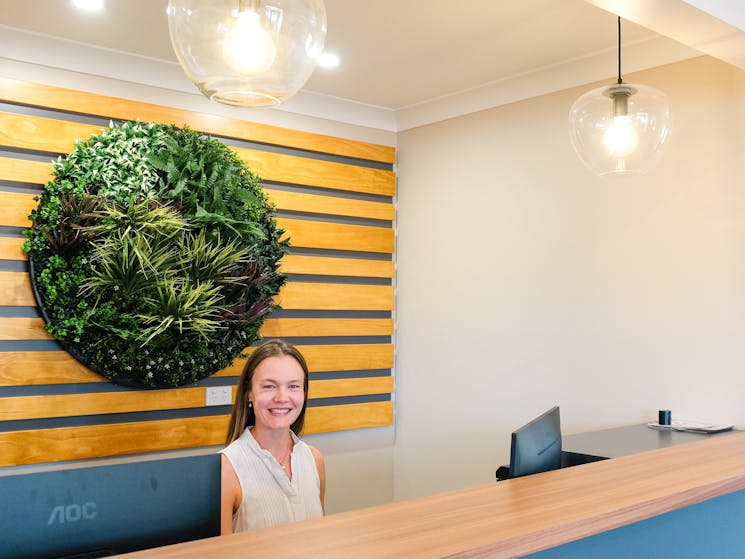 Inside reception area with a women sitting behind the desk. Green round plant in the background