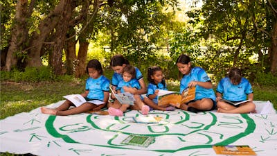 A group of diverse young children are sitting outdoors under lush green trees, engaged in reading.