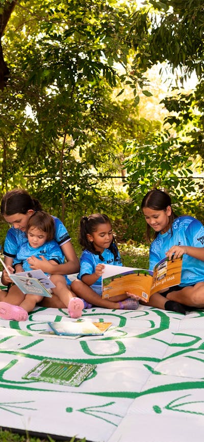 A group of diverse young children are sitting outdoors under lush green trees, engaged in reading.