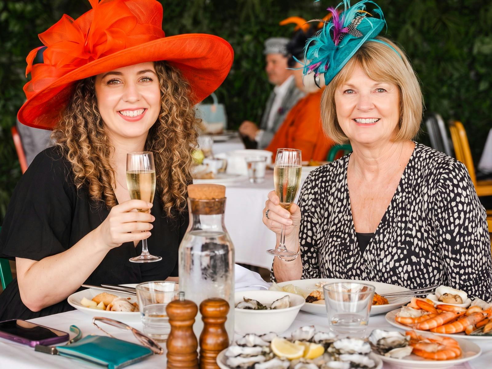 Two guests wearing race day hats enjoying drinks and dining at a Melbourne Cup event.