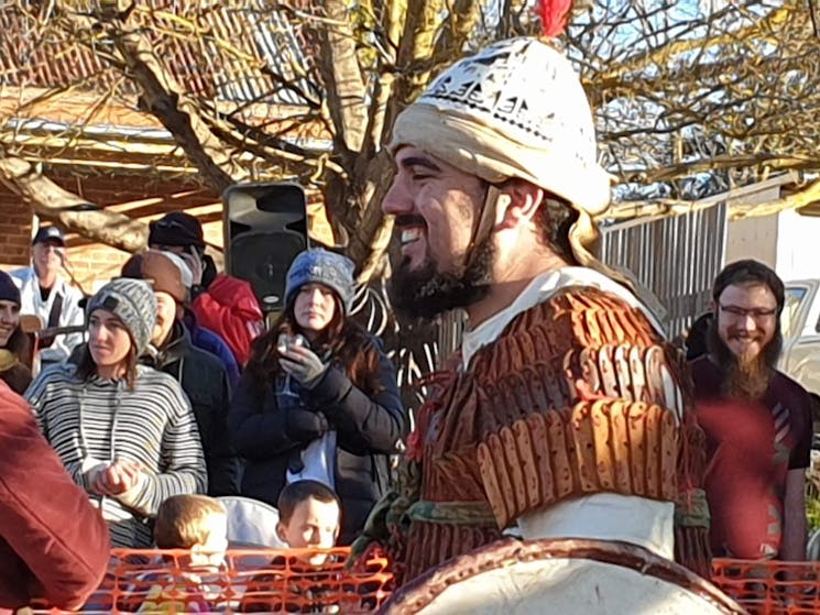 Upper body view of a bearded man dressed in Viking armour and helmet; small crowd in background
