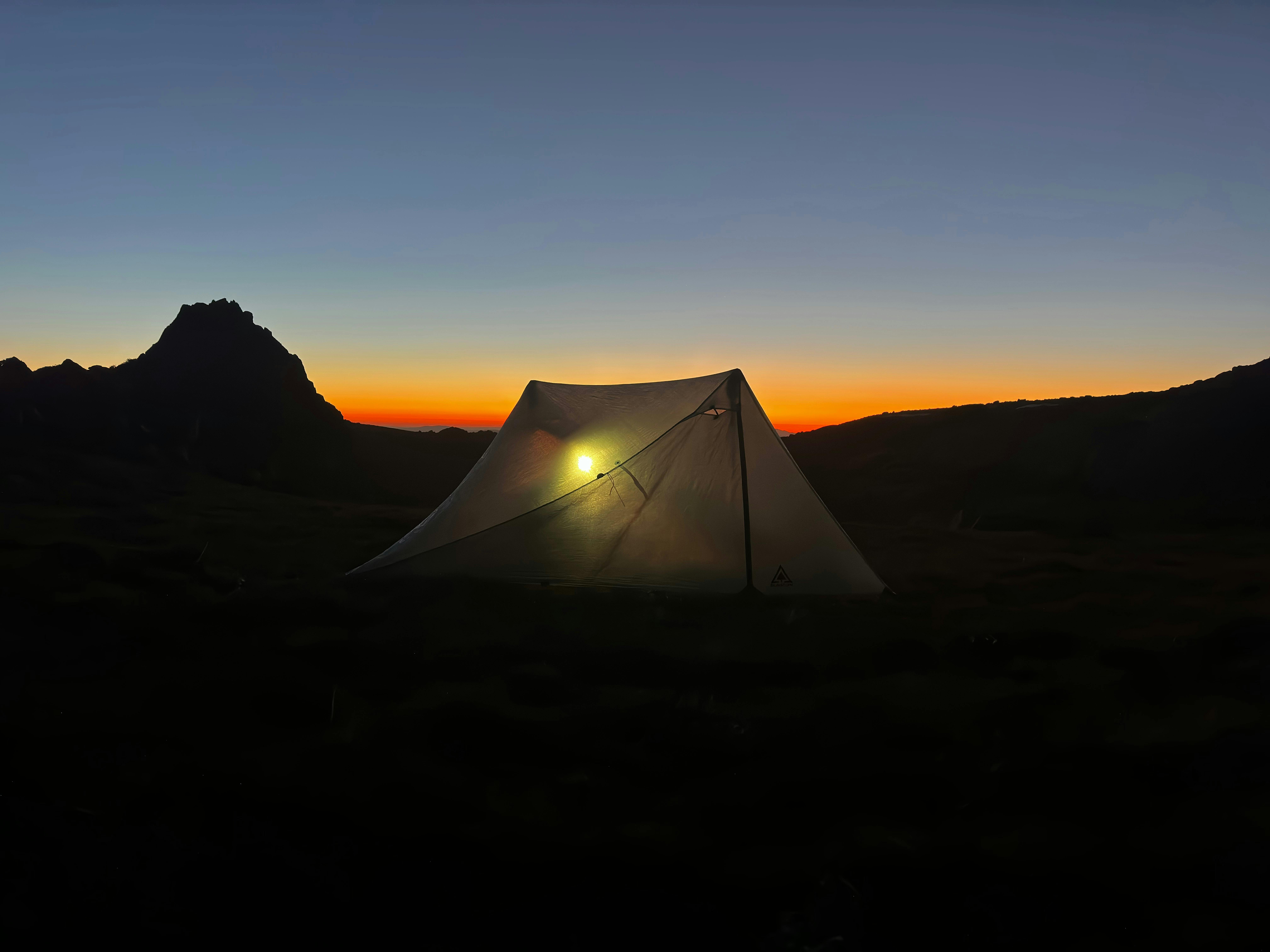 A tent in the mountains with the sun setting behind it.