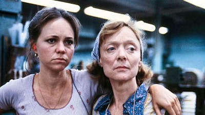 Two women look tired, one has arm over the other both looking to their left towards something
