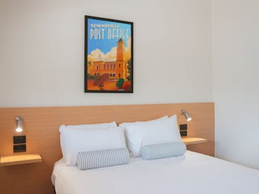 Queen bed in a bright hotel room with reading lights and framed Townsville Post Office artwork.