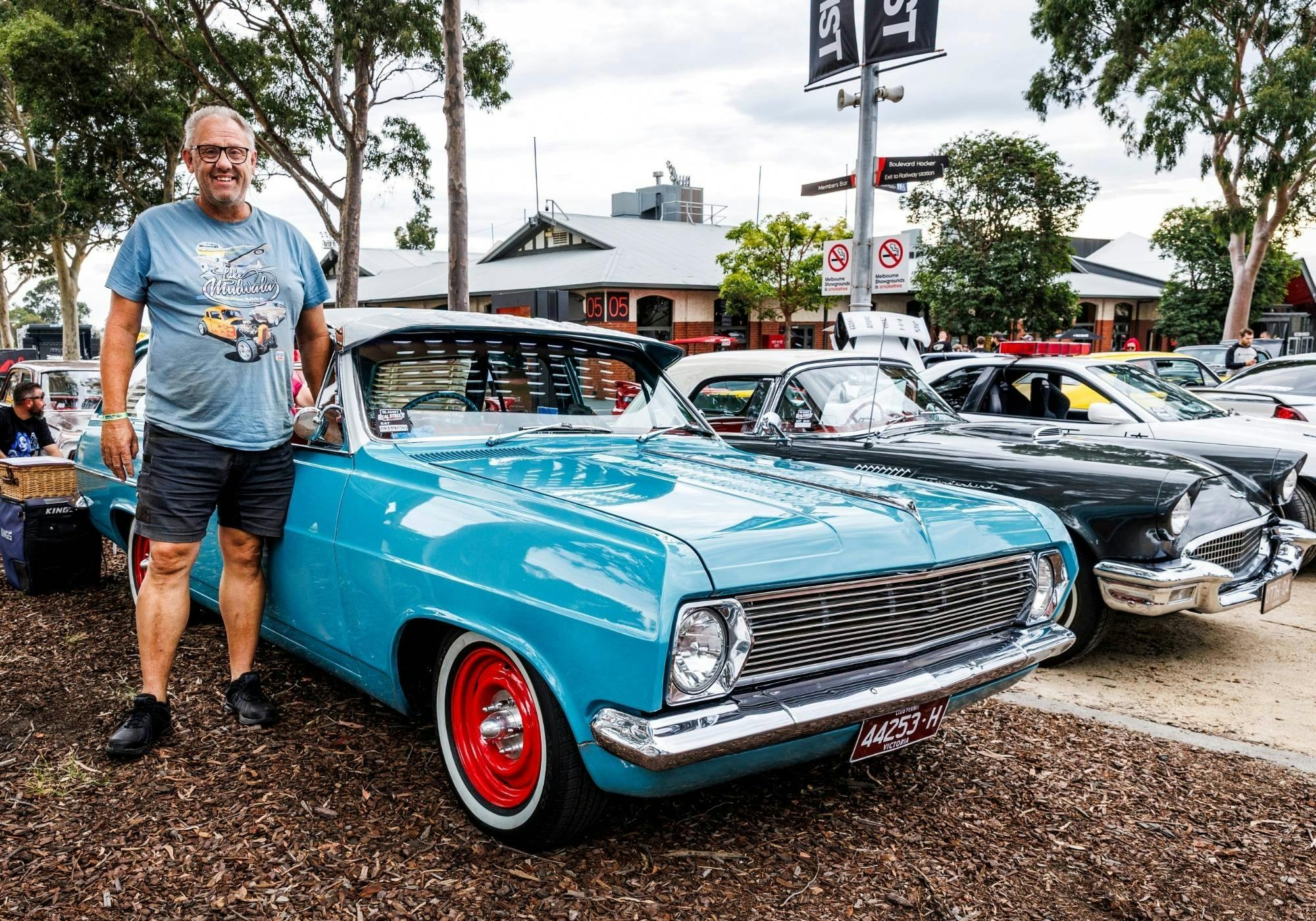 Smiling owner standing beside a turquoise classic car with red wheels at an outdoor display.