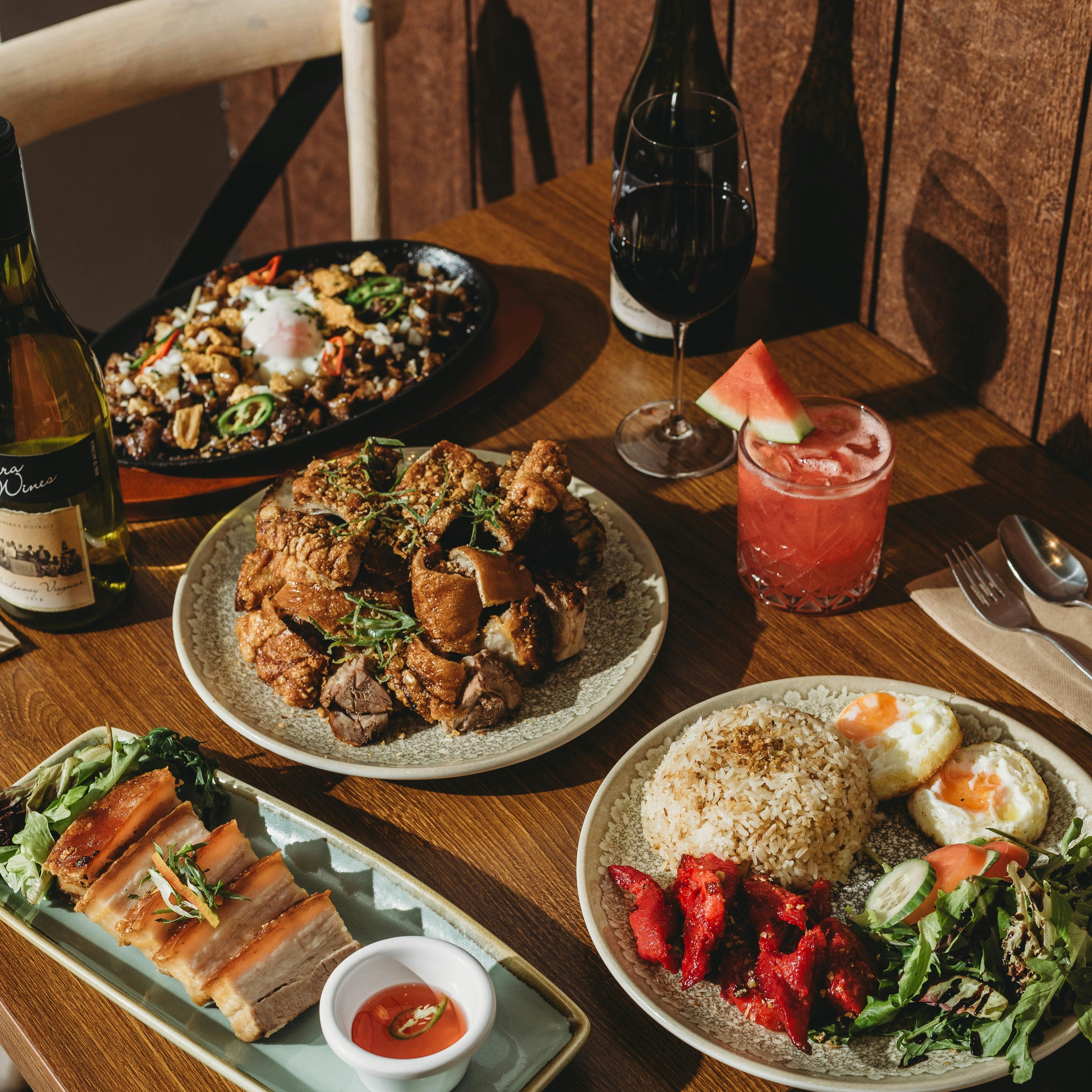 A wooden dining table filled with Filipino dishes including crispy pork belly, fried rice with eggs,