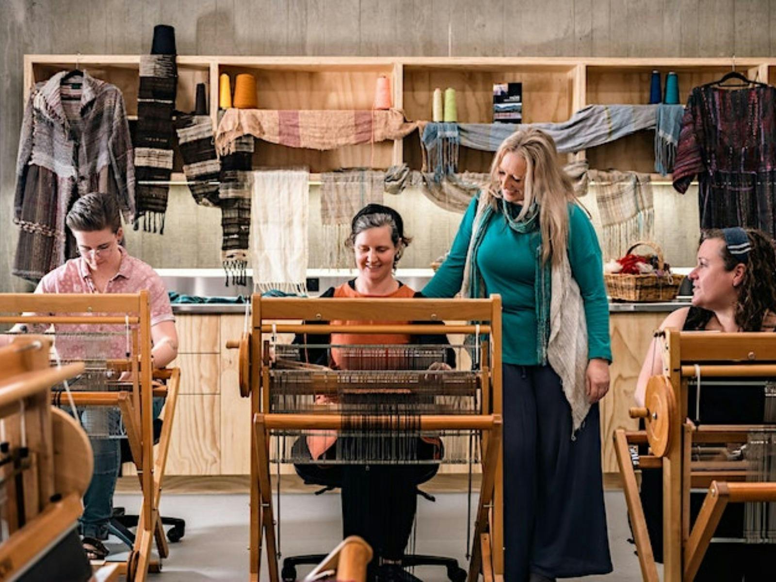 Three women seated at looms with instructor standing between them inside workshop