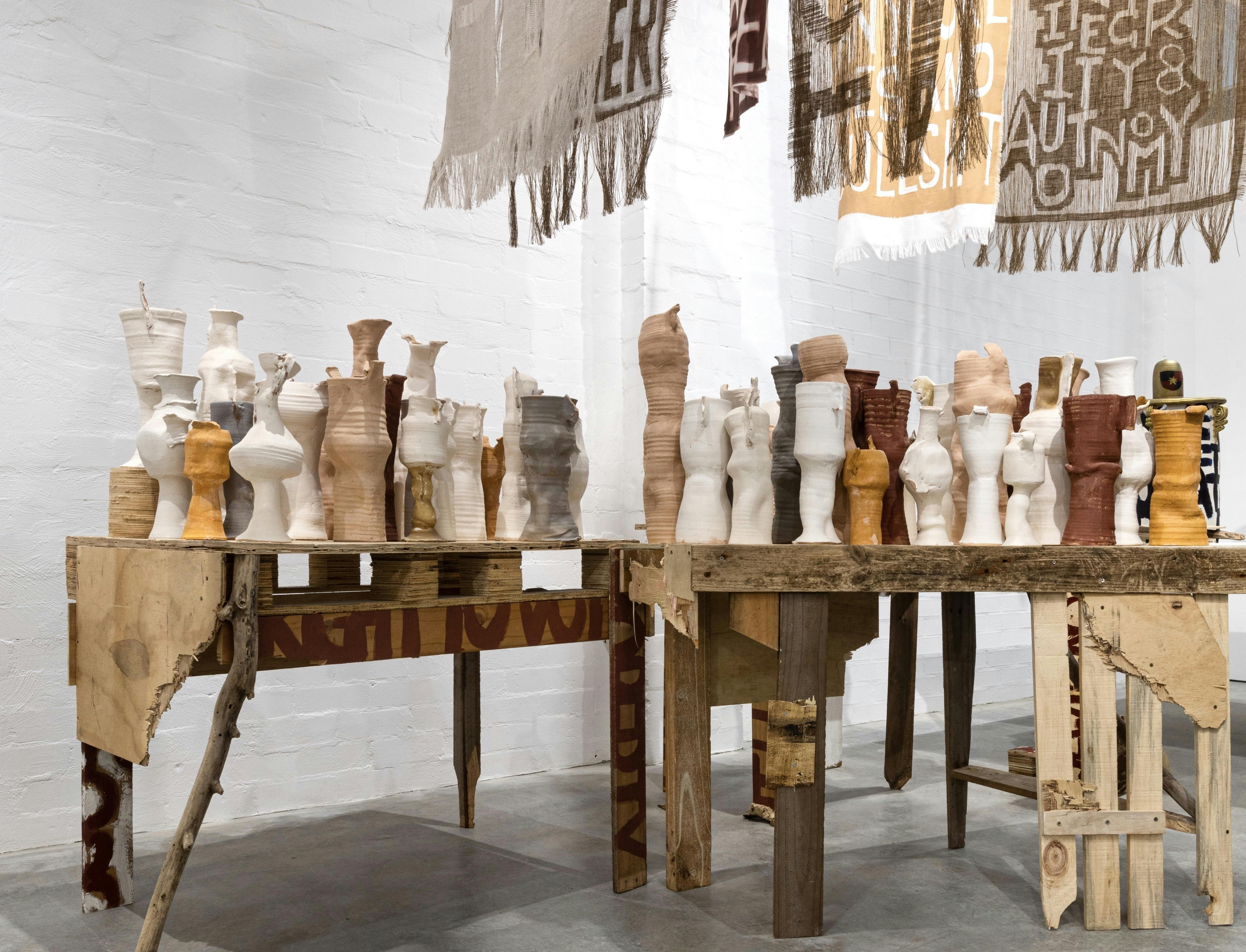 Raw clay vessels on timber tables beneath protest banners in Kara Wood’s Enough installation.