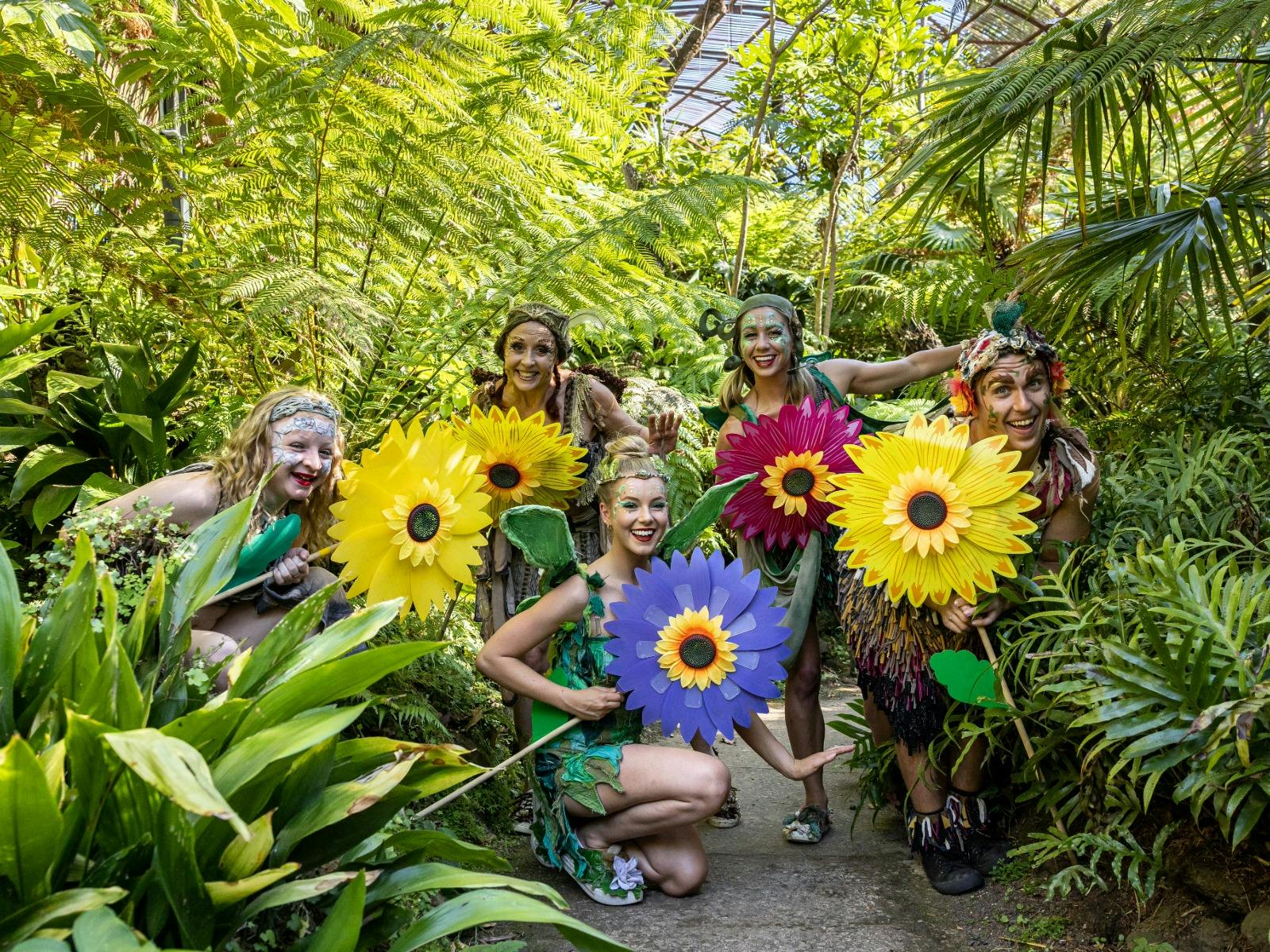 Five people dressed as fairies holding large flowers, standing in a vibrant garden.