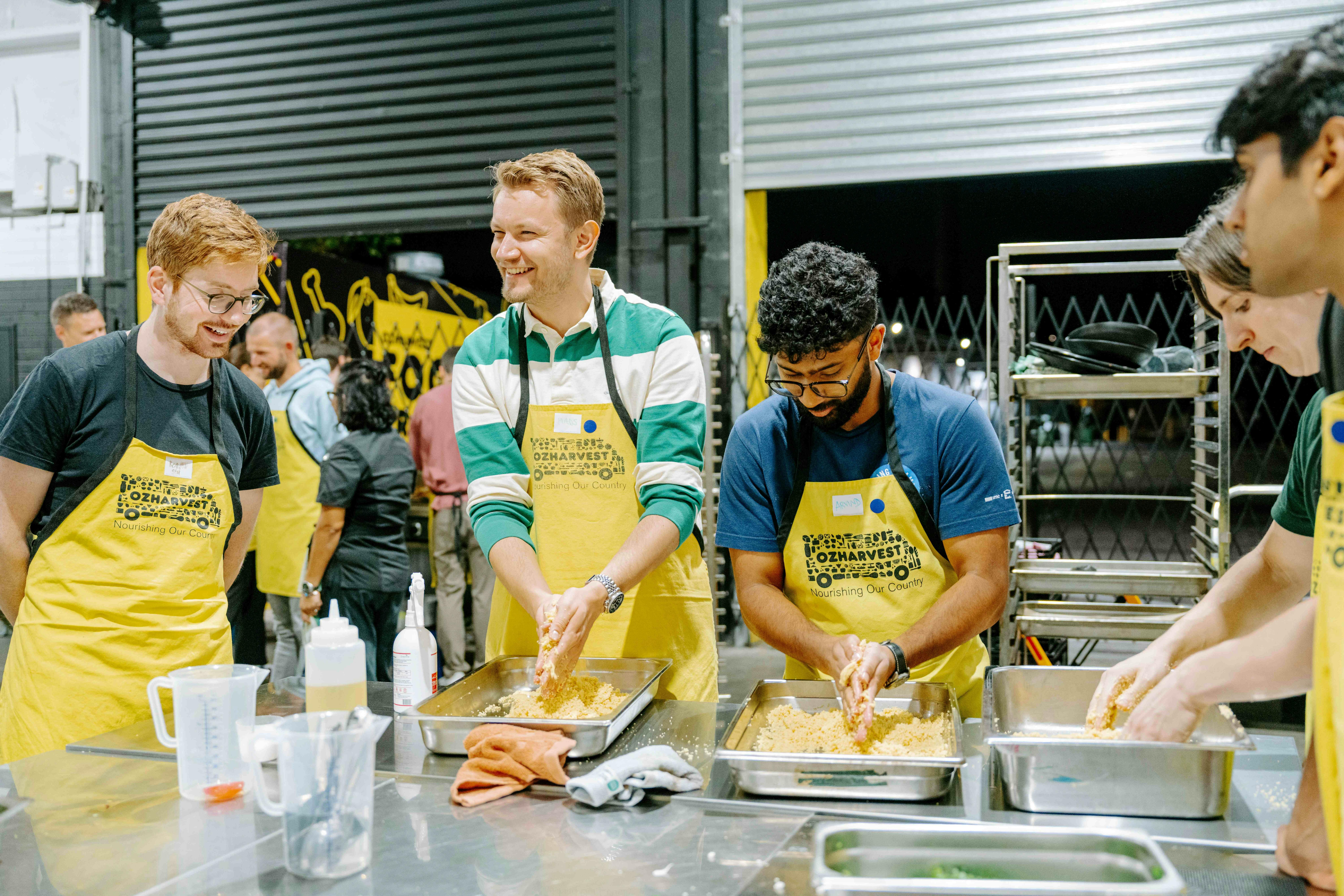 A group of people wearing OzHarvest aprons mixing and shaping food by hand in metal trays, in a ware