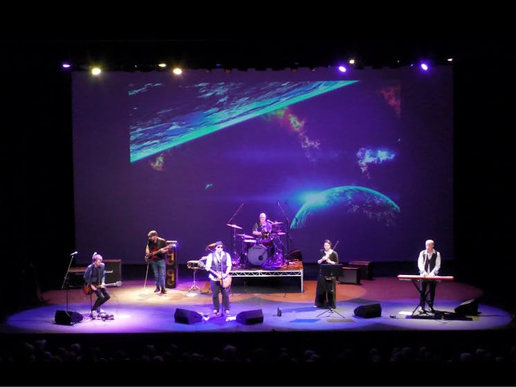 A group of people on stage with various instruments performing in front of a screen with projections