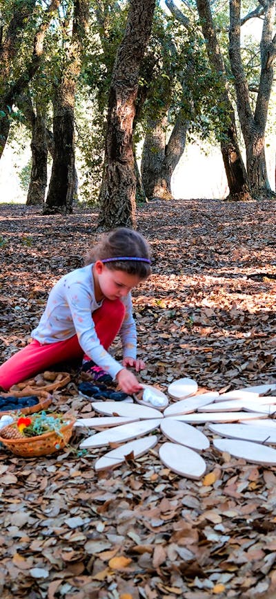 A young girl plays with wooden mandala shapes on a leafy forest floor