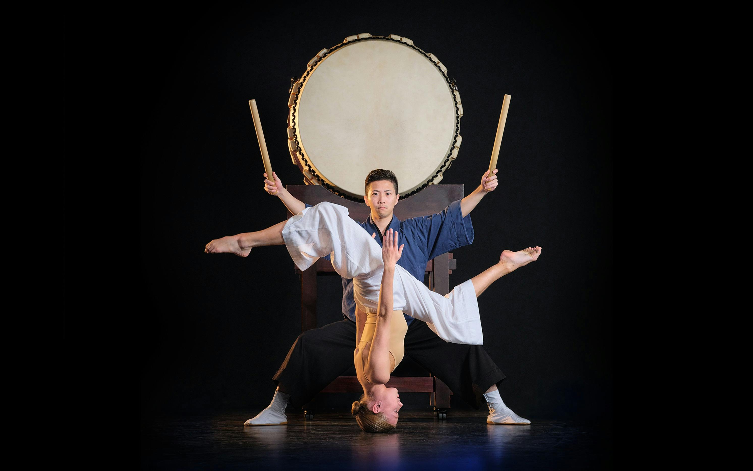 Acrobat balances upside down before a taiko drummer holding sticks in a dramatic pose.
