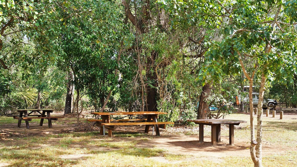 Picnic table under shade with vehicles and camping equipment in the background.