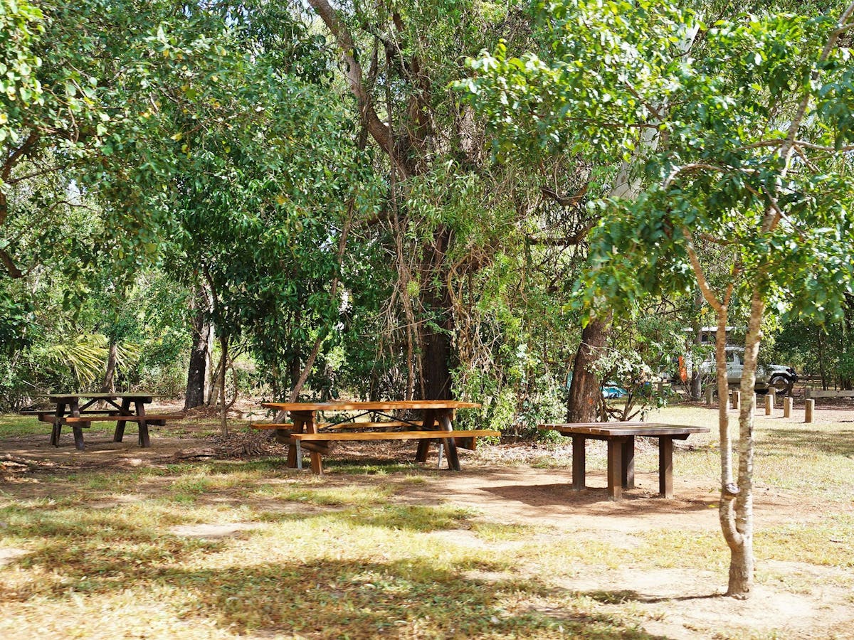 Picnic table under shade with vehicles and camping equipment in the background.