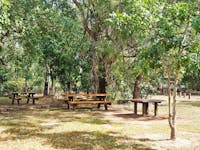 Picnic table under shade with vehicles and camping equipment in the background.