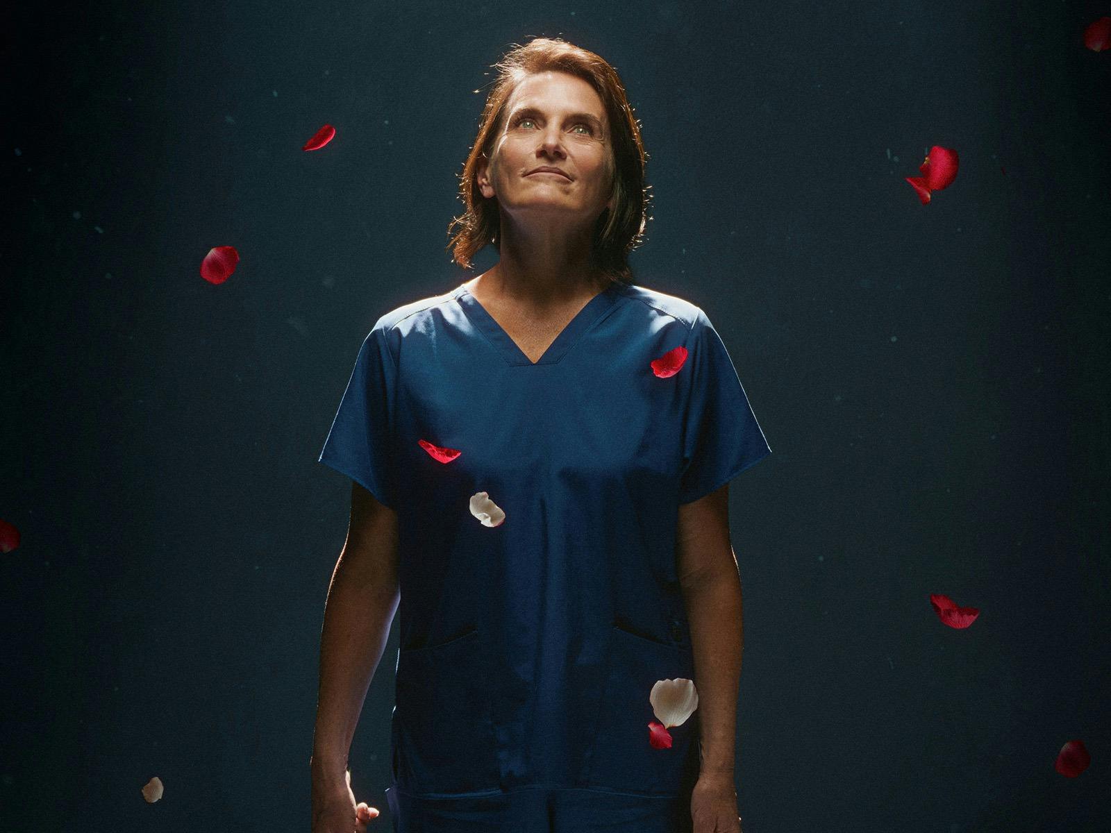 A middle aged woman wearing scrubs holds a bunch of rosed as petals fall from the sky.