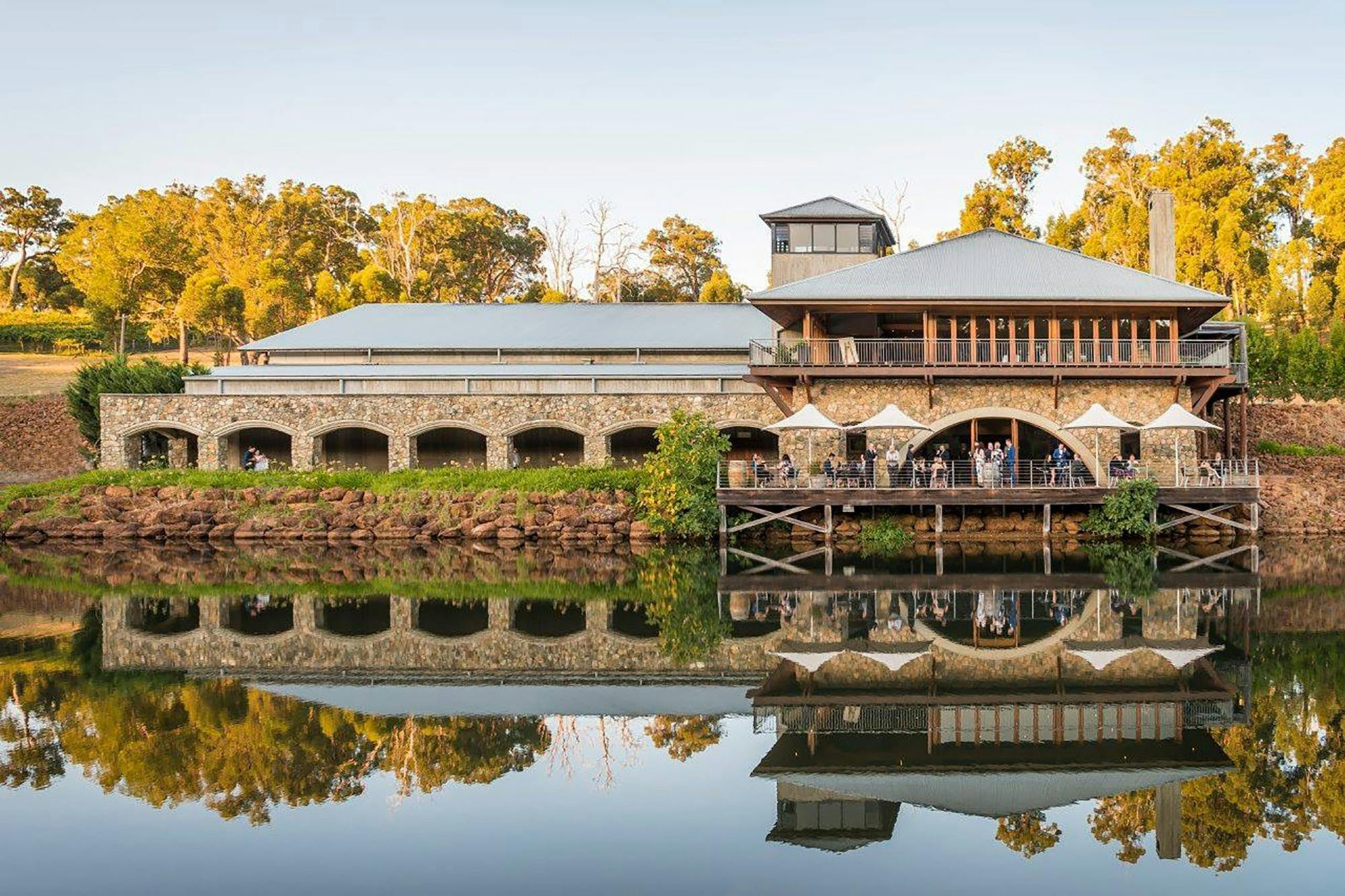 A large building with stone and wood elements is reflected in a calm body of water situated infront