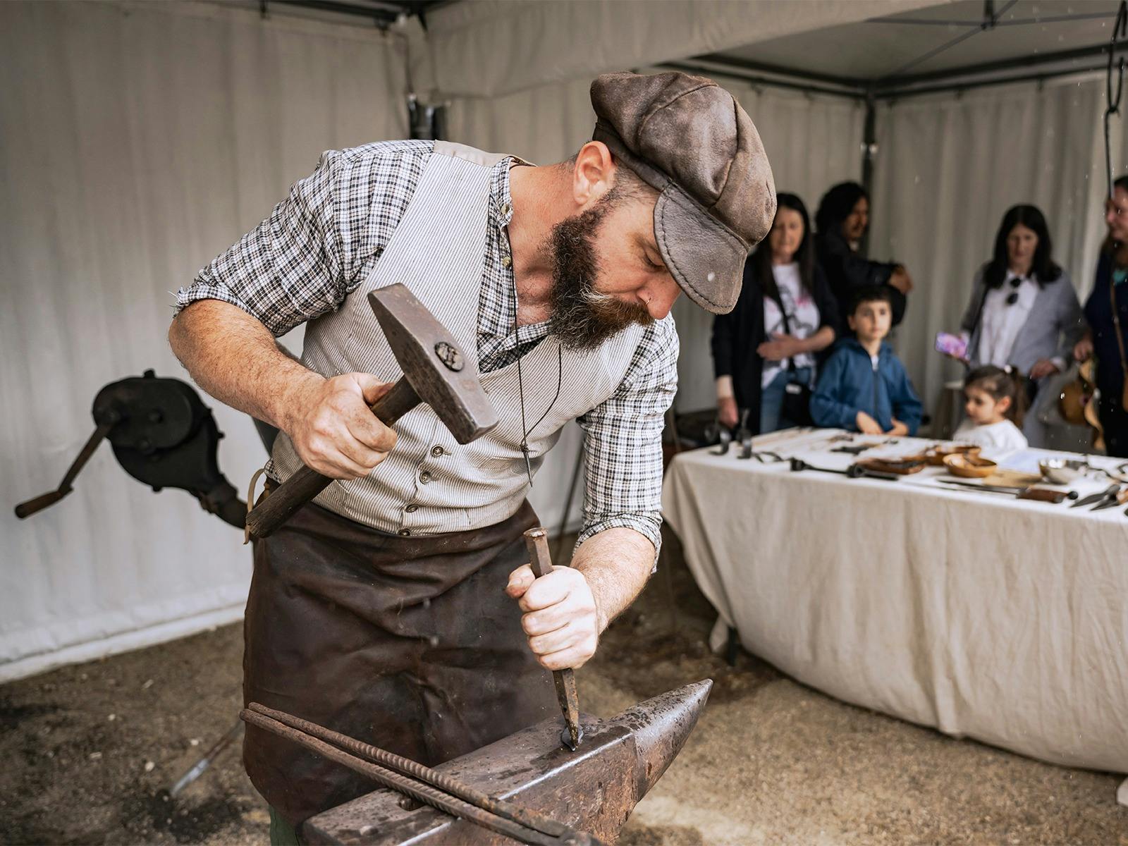 A blacksmith in vintage attire skillfully hammers metal on an anvil, watched by an engaged audience.