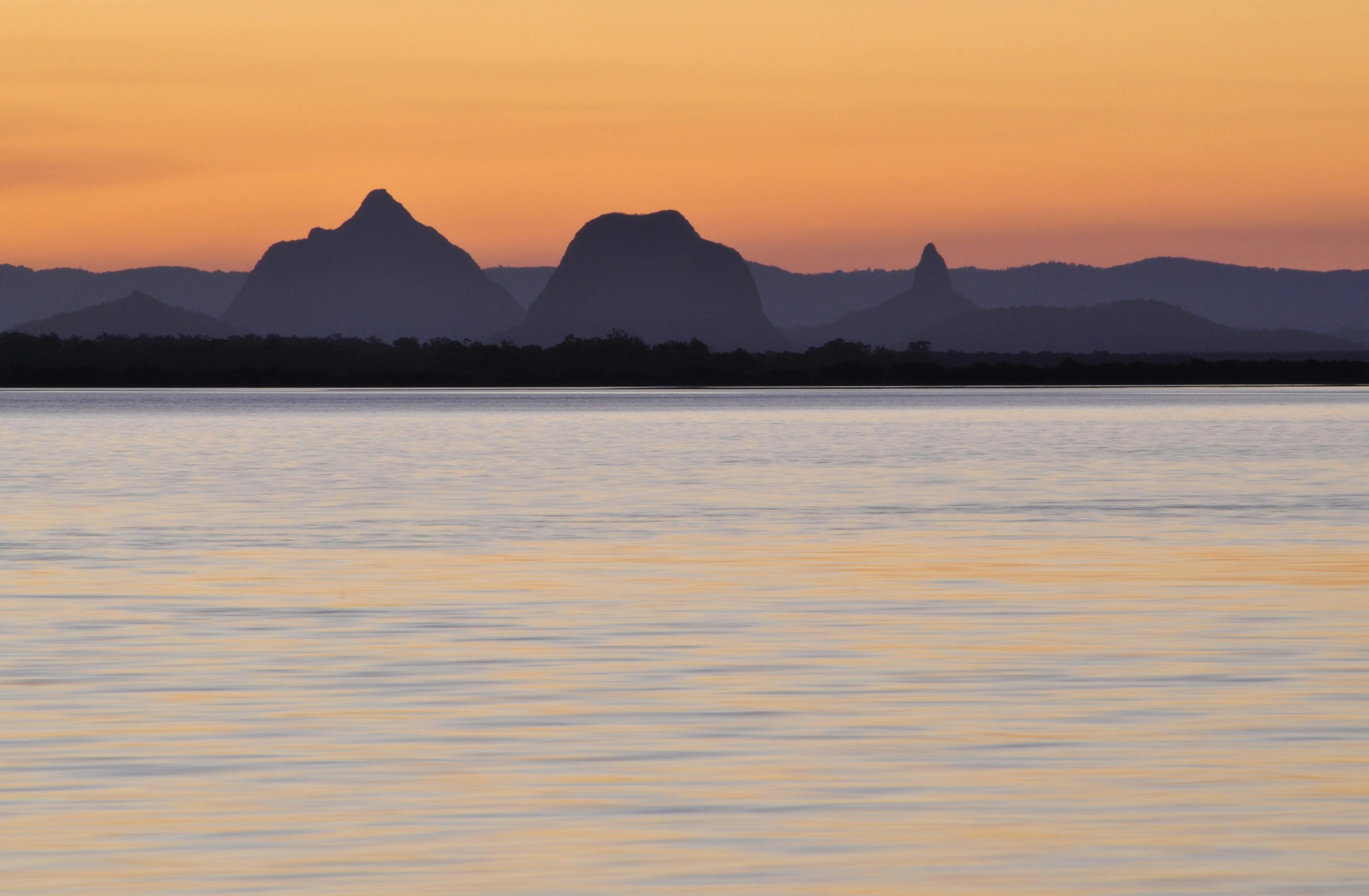 Sunrise view over calm water with the silhouetted peaks of the Glass House Mountains