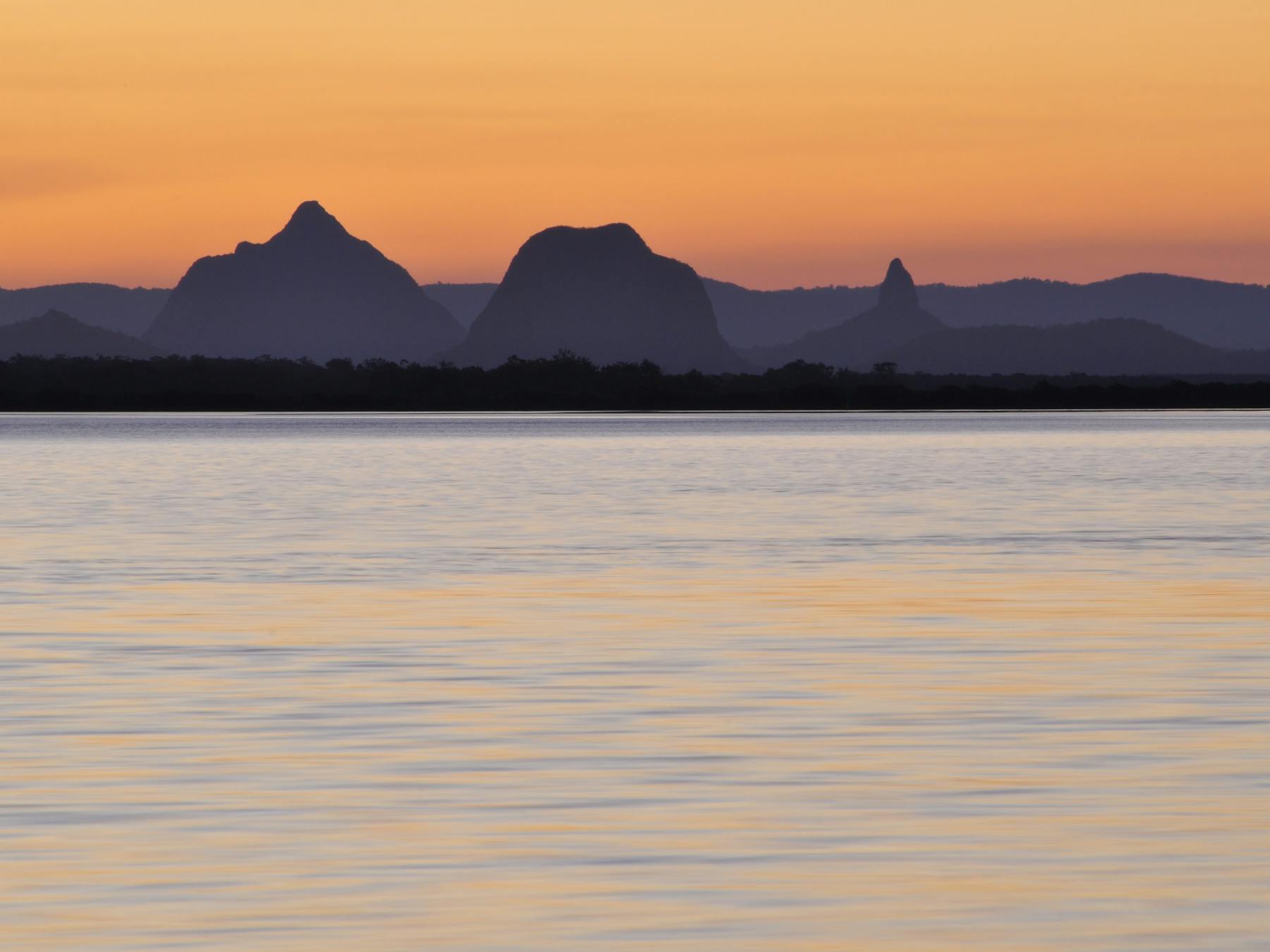Sunrise view over calm water with the silhouetted peaks of the Glass House Mountains