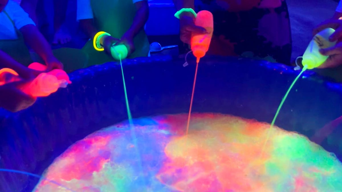 Four Messy Glow Co participants pouring glowing paint into a pool together making a glowing rainbow