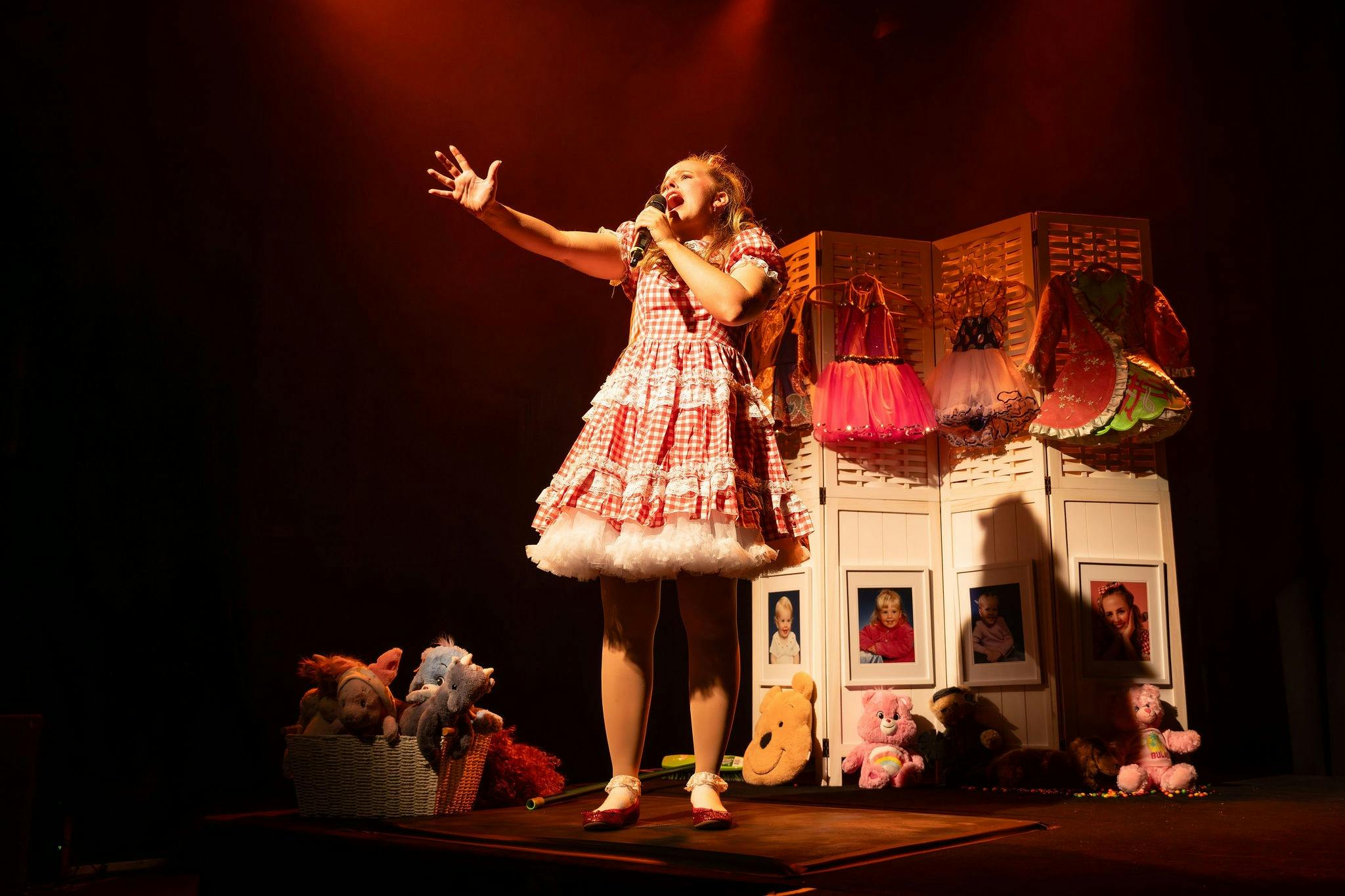 A performer in a red gingham dress sings into a microphone onstage, reaching into audience