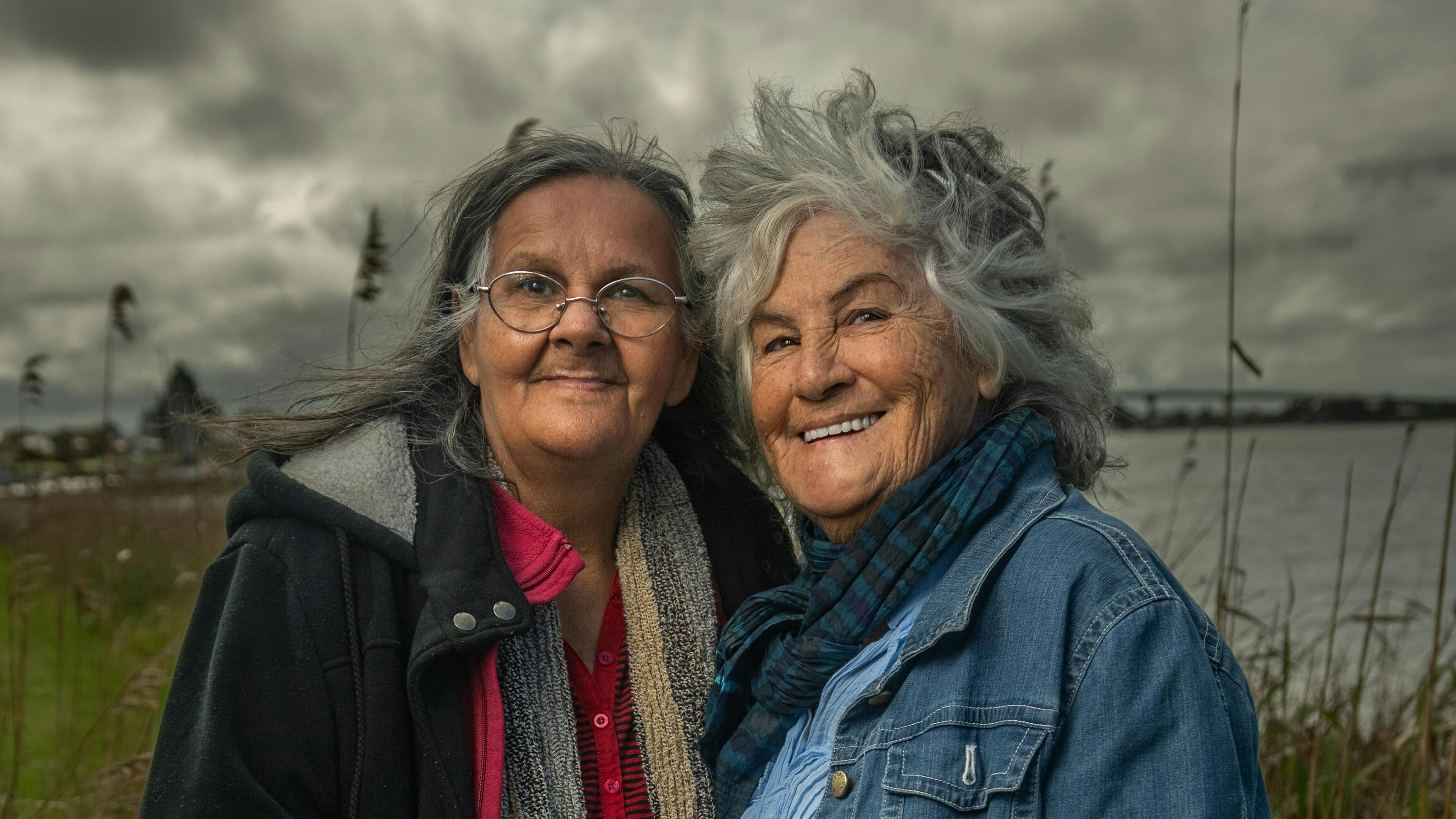 Two First Nations female Elders in front of water and a moody sky.