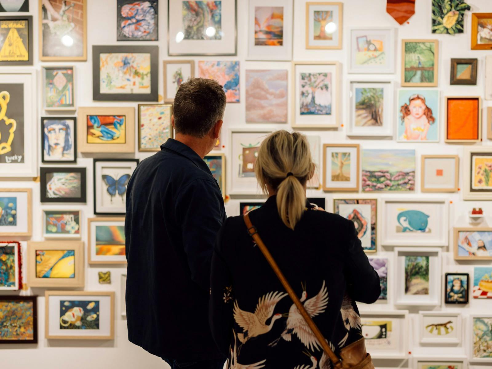 Two people stand looking at a gallery wall, filled with framed artwork.