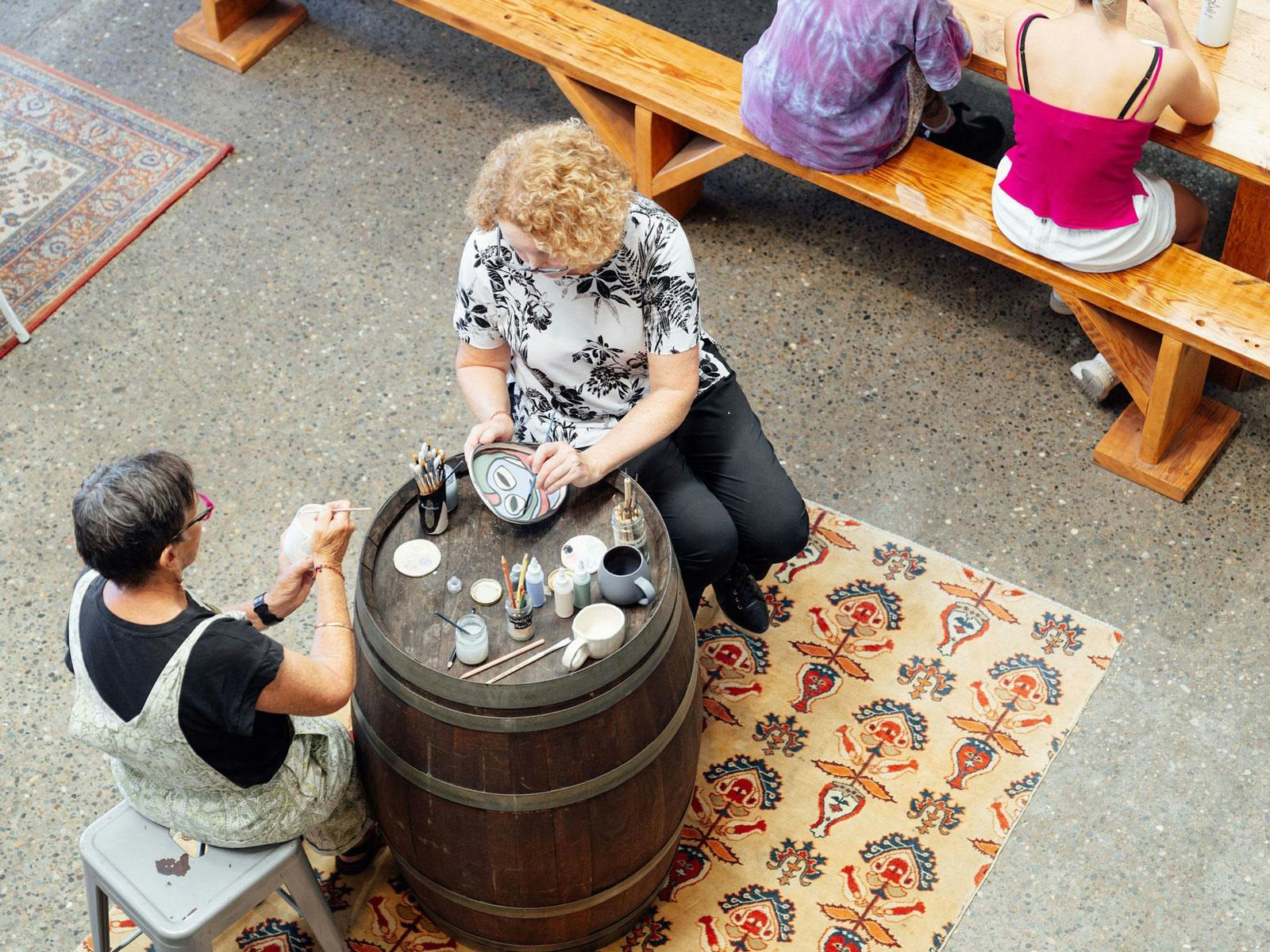 2 people painting bisque fired pieces whilst sitting on bar stools at a wine barrel