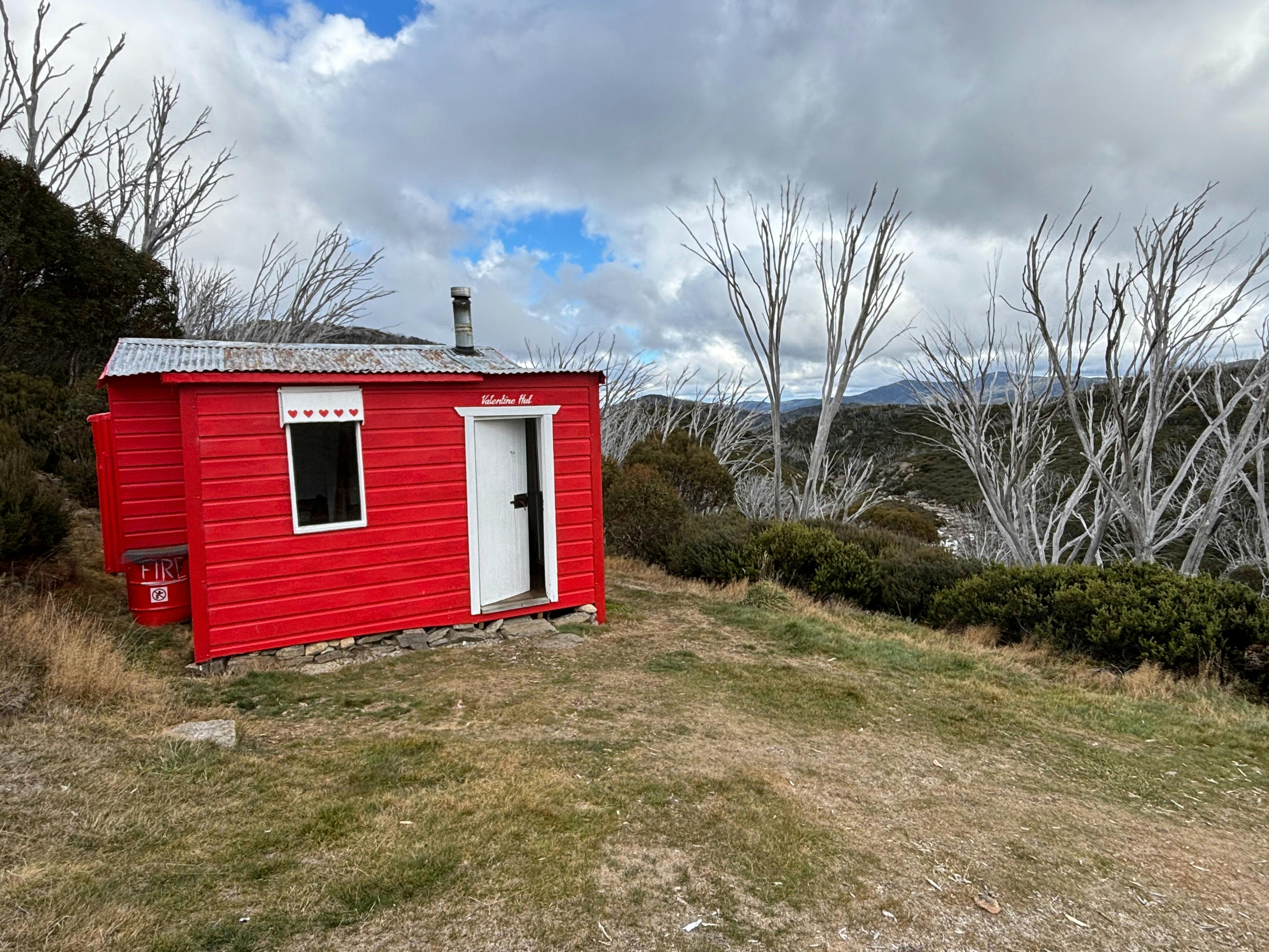 A bright red timber hut on the side of a hill with burnt snow gum trees behind.