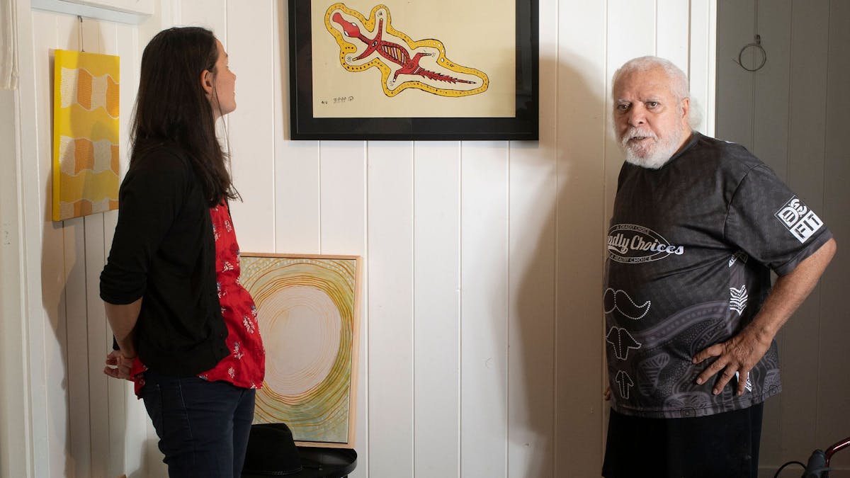 Two people standing in front of art, one older man facing the camera with white hair and beard
